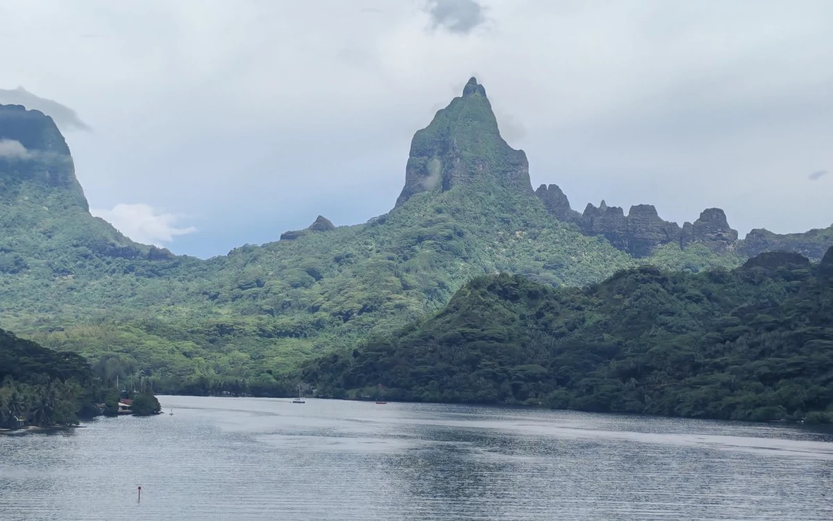 A long bay between green slopes, with a jagged rock peak rising above it under cloud