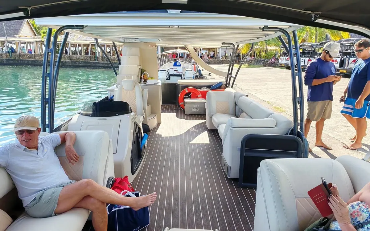 White padded sofas and armchairs under a canopy on a striped wooden deck, moored by a quay with palm trees